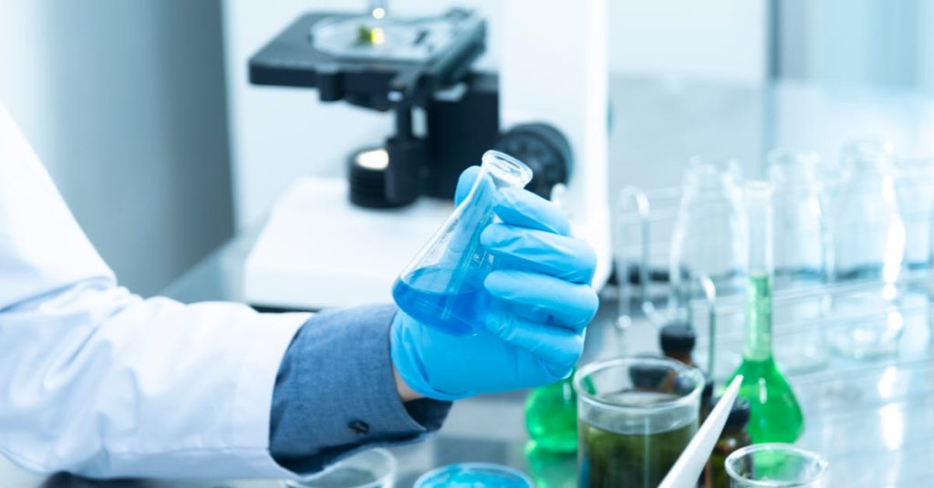 Scientist in gloves analyzing blue liquid in a laboratory setting with microscope and glassware.